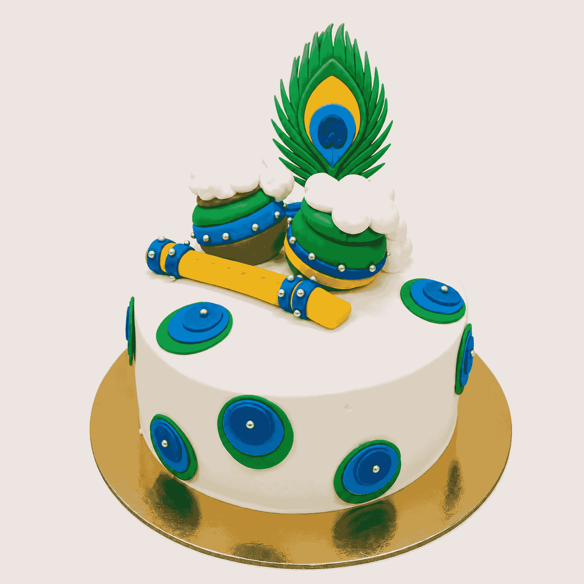 A round white festive cake decorated with layered peacock feather medallions, topped with two butter pots spilling white cream, a yellow flute, and a peacock feather.