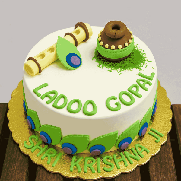 A round white festive cake decorated with peacock feather cutouts around the base, topped with a clay pot on green grass, a yellow flute, and green Ladoo Gopal script.