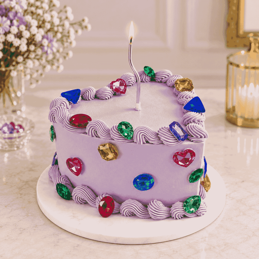 A heart-shaped lavender celebration cake with piped shell borders, colorful faceted gemstone crystals, and a slender silver celebration candle in the center.