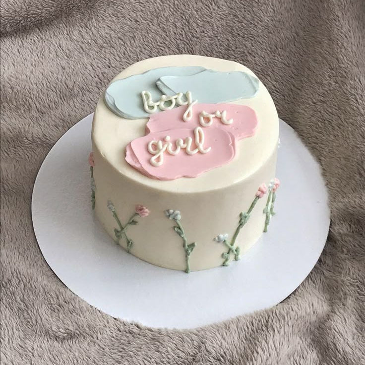 A minimalist white cake with a painterly blue and pink frosting stroke design on top, cursive writing saying boy or girl, and small piped wildflowers on the sides.
