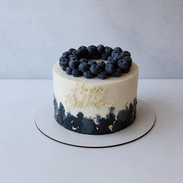 A premium round birthday cake for him featuring a smooth ivory top, a textured black brushstroke base with gold leaf flakes, a gold Happy Birthday script topper, and a crown of fresh blueberries.