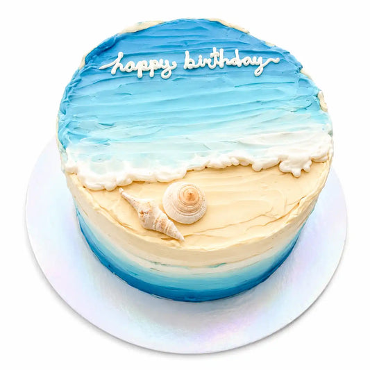 A beautiful round beach themed birthday cake designed with palette-knife textures showing sandy cream shorelines, a frothy white wave, blue ocean water, and hand-molded chocolate seashells as seen in image.