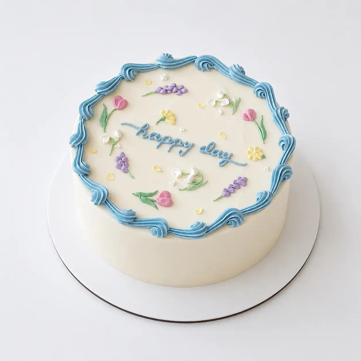 A minimalist white round Korean style cake decorated with a wavy blue buttercream border, small hand-piped pastel flowers like tulips and lavender, and blue cursive writing reading happy day.
