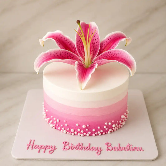 A tall round birthday cake featuring a smooth white to vibrant pink ombré frosting gradient, decorated with a border of pink and white sugar pearls around the base, custom script lettering on the board, and a large pink tiger lily bloom on top as seen in image.