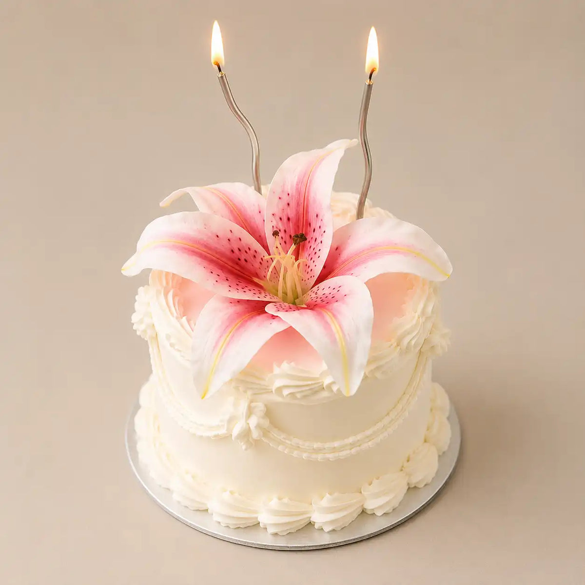 An elegant tall premium white celebration cake decorated with vintage cream ruffles and a large pink tiger lily on top, accompanied by two lit wavy silver candles inside a pink illustrated box with bow designs.