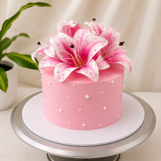 A tall round premium pastel pink birthday cake on a metallic rotating stand, decorated with tiny scattered white pearls and topped with a large, dense crown of pink and white stargazer lilies covered in glistening edible dewdrops as seen in image.