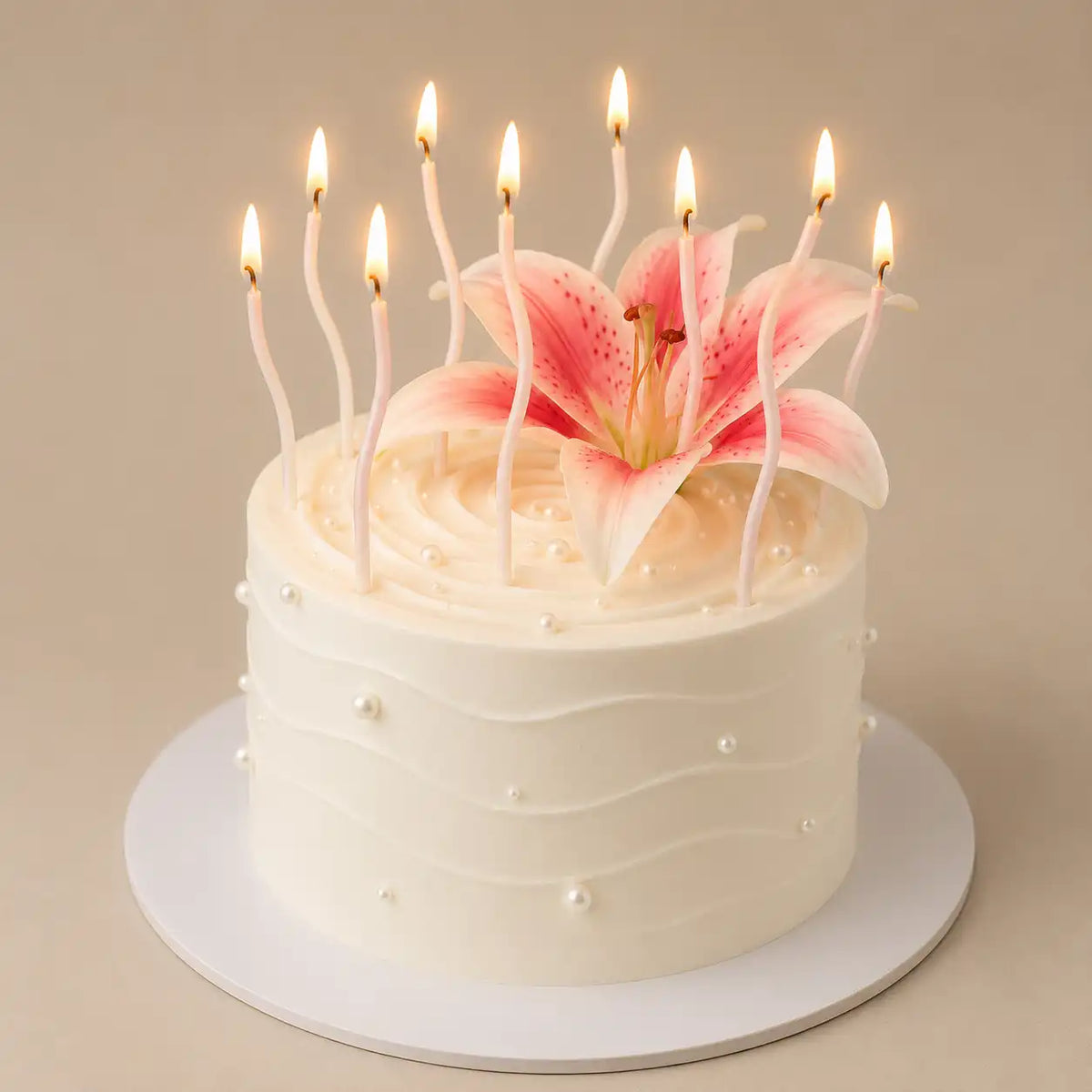 A tall round premium white celebration cake with horizontal rippled icing and tiny white pearls, featuring a large open pink lily in the center surrounded by multiple lit, slender white wavy candles.
