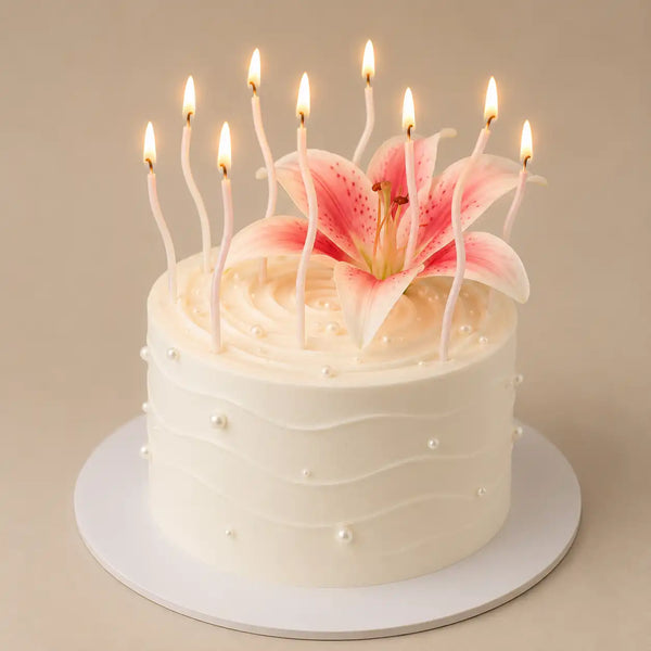 A tall round premium white celebration cake with horizontal rippled icing and tiny white pearls, featuring a large open pink lily in the center surrounded by multiple lit, slender white wavy candles.
