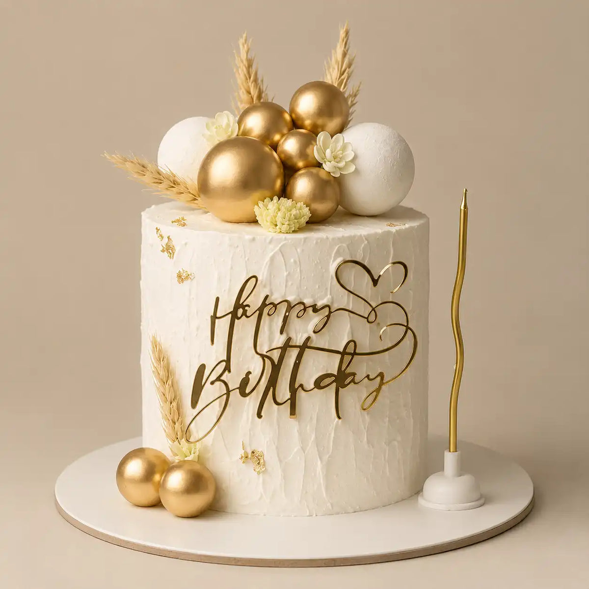 A tall round premium birthday cake with a heavily textured white vertical cream body decorated with gold leaf flakes, topped with white and polished gold chocolate spheres, dried wheat stalks, an elegant gold Happy Birthday script topper, and a wavy gold candle as seen in image.