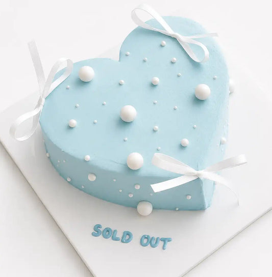 A minimalist heart-shaped light blue cake decorated with scattered small and large white edible pearls and three dainty hand-tied white satin ribbons around the edges as seen in image.