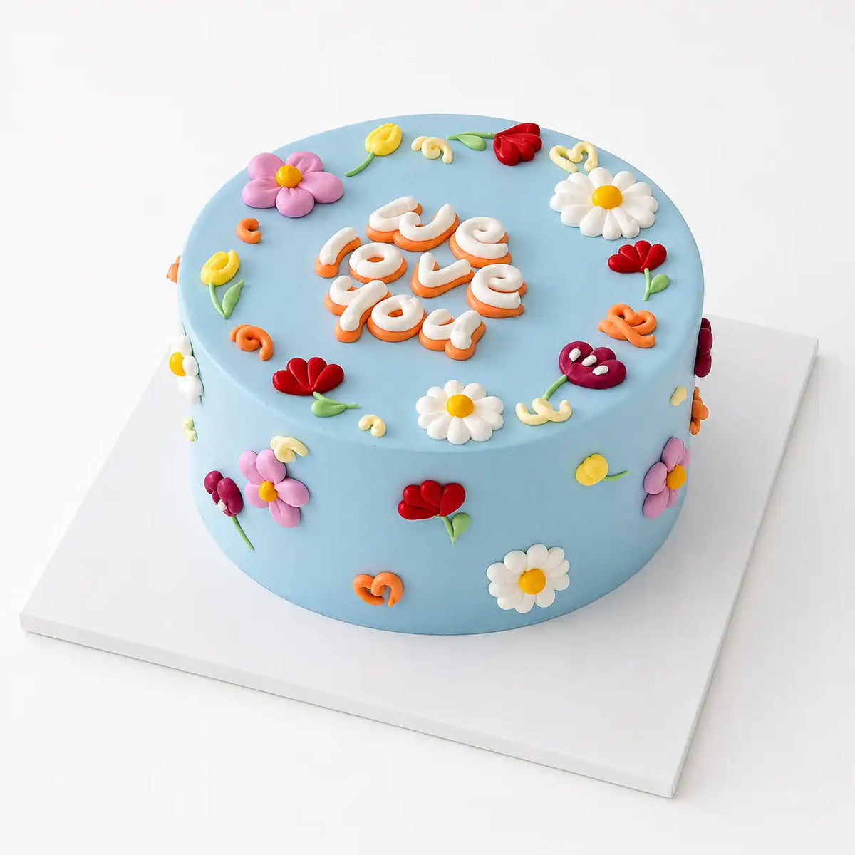 A minimalist light blue round Korean style cake decorated with a scatter of hand-piped white daisies, red tulips, and purple flowers, featuring a chunky 3D text message reading we love you in the center as seen in image