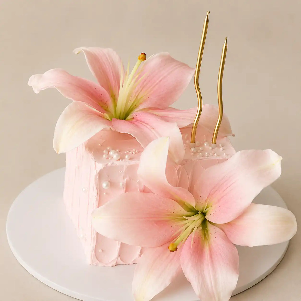 A premium square celebration cake with light pink textured frosting and scattered white pearls, featuring one large pink lily on top, another at the base, and two tall wavy gold candles as seen in image.