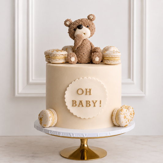 A premium smooth ivory baby shower cake decorated with gold-flecked macarons and an 'OH BABY!' plaque, topped with a detailed, textured brown edible teddy bear.