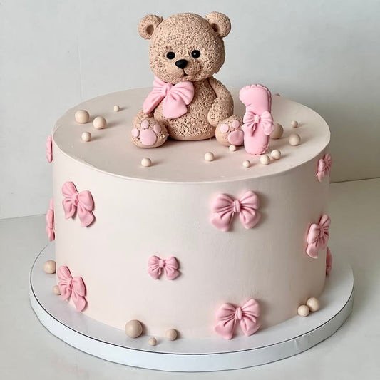 A smooth light pink baby shower cake decorated with small pink fondant bows and chocolate pearls, topped with an intricately textured brown edible teddy bear wearing a pink bow.