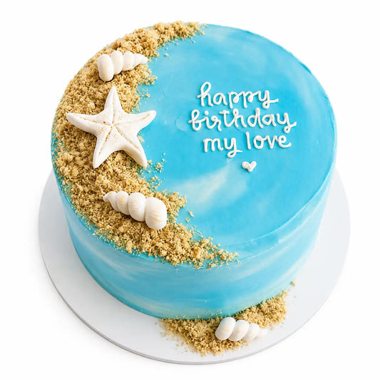 A minimalist round turquoise and white watercolor birthday cake decorated with a crescent of cookie crumble sand, a chocolate starfish, seashells, and white hand-piped cursive writing reading happy birthday my love as seen in image.