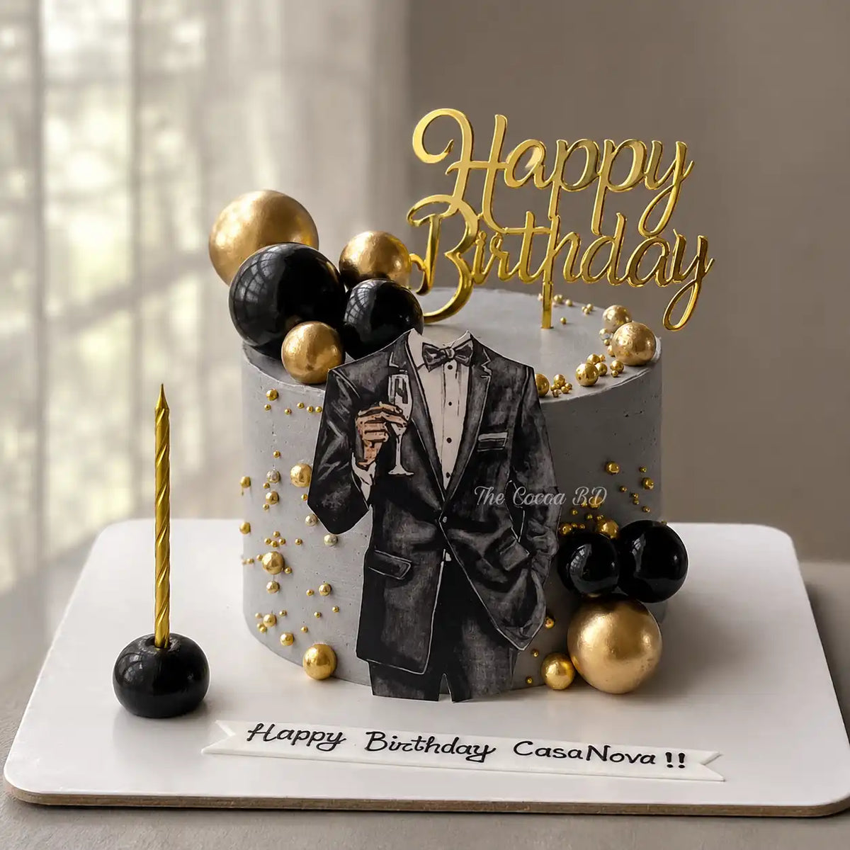 A tall round charcoal-grey birthday cake for him featuring a tuxedo silhouette cutout holding a champagne glass, decorated with high-gloss black and gold spheres and a gold Happy Birthday topper as seen in image
