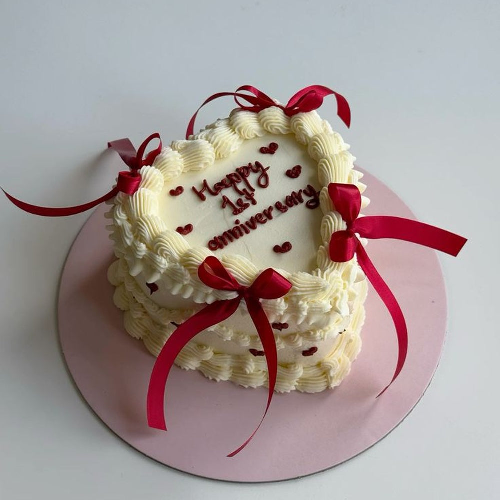 An elegant heart-shaped white vintage cake with detailed ruffle piping, decorated with real red satin ribbon bows, tiny piped red hearts, and 'Happy 1st anniversary' in red script.