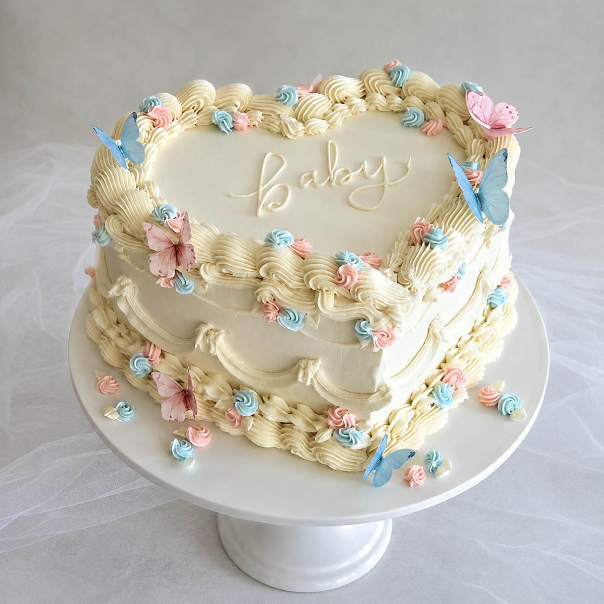 An elegant heart-shaped ivory vintage cake with detailed ruffle piping, decorated with pastel pink and blue butterflies and the word baby written in cursive.