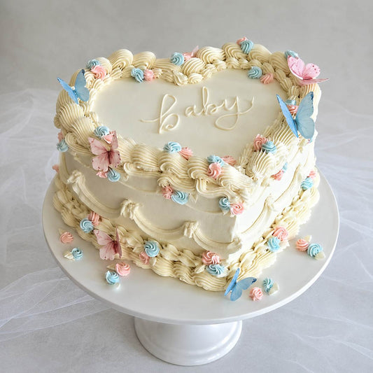 An elegant heart-shaped ivory vintage cake with detailed ruffle piping, decorated with pastel pink and blue butterflies and the word baby written in cursive.